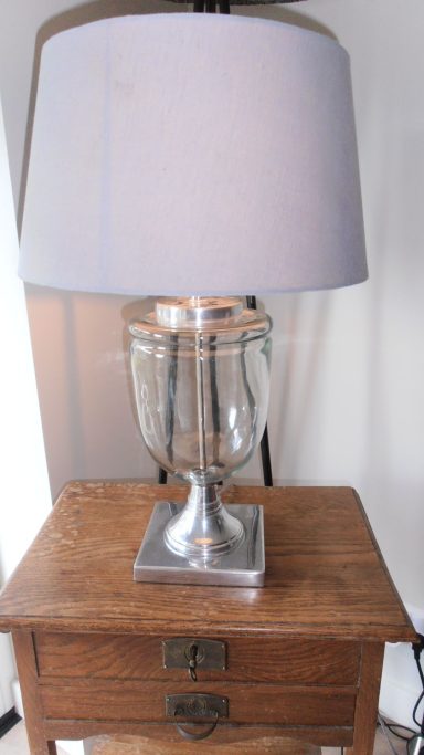 Glazen tafellamp met een grijze lampenkap op een houten nachtkastje.