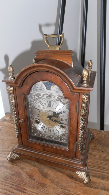 Antieke houten klok met metalen details en wijzerplaat, op een houten tafel.