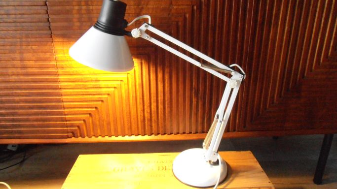 Witte bureau lamp met verstelbare arm en houten ondergrond, afgezet tegen een houten wand.